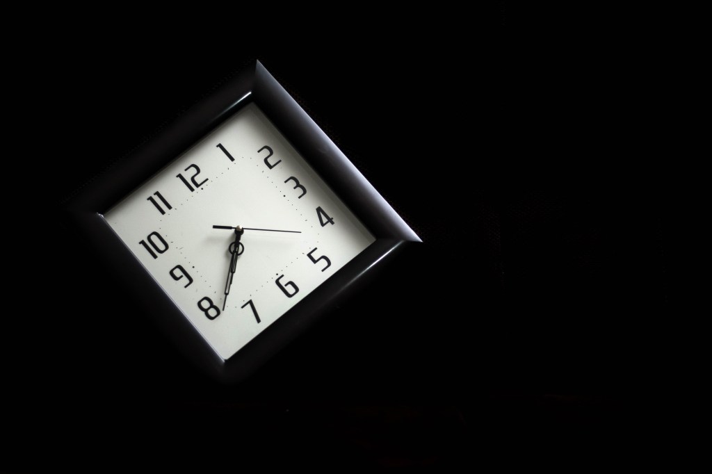 A black clock with a white face hangs diagonally in darkness. The hands read thirty eight past seven o'clock.